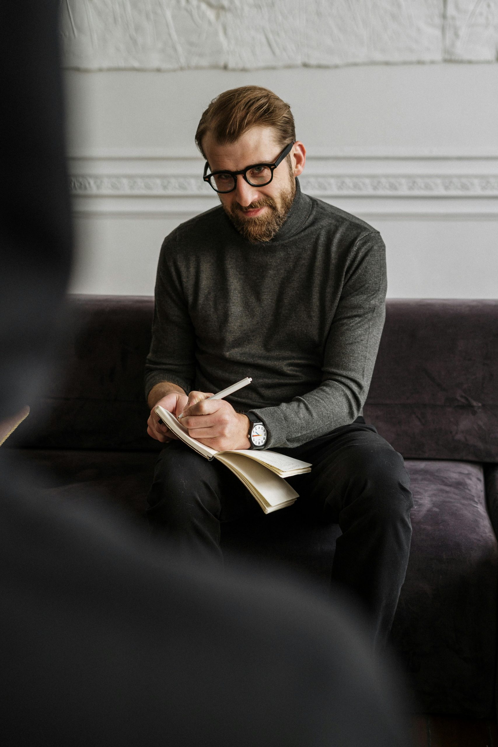 Man in gray sweater and black pants sitting on purple couch